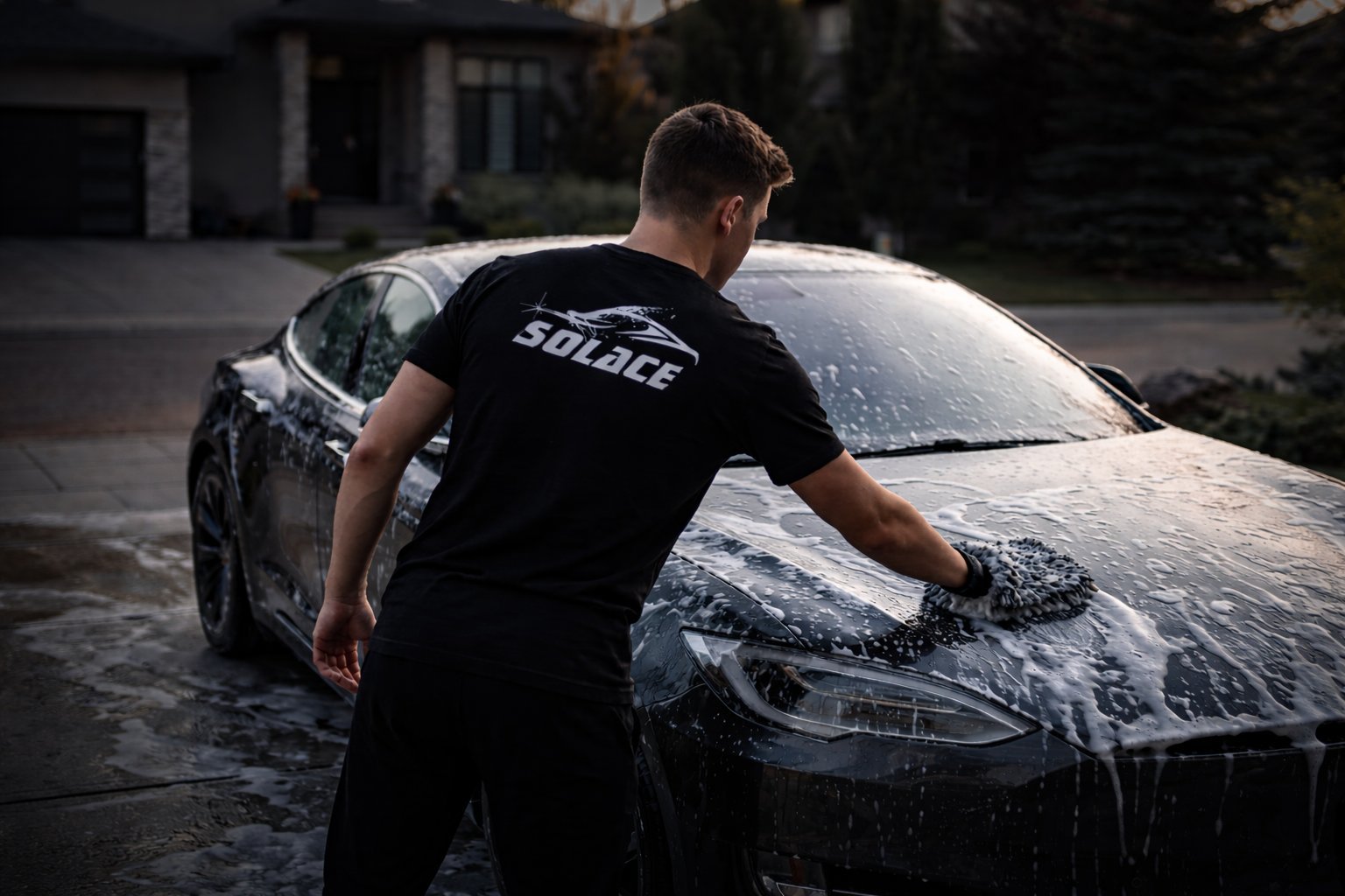 Man in black t-shirt washing a dark car with soapy water in a residential driveway