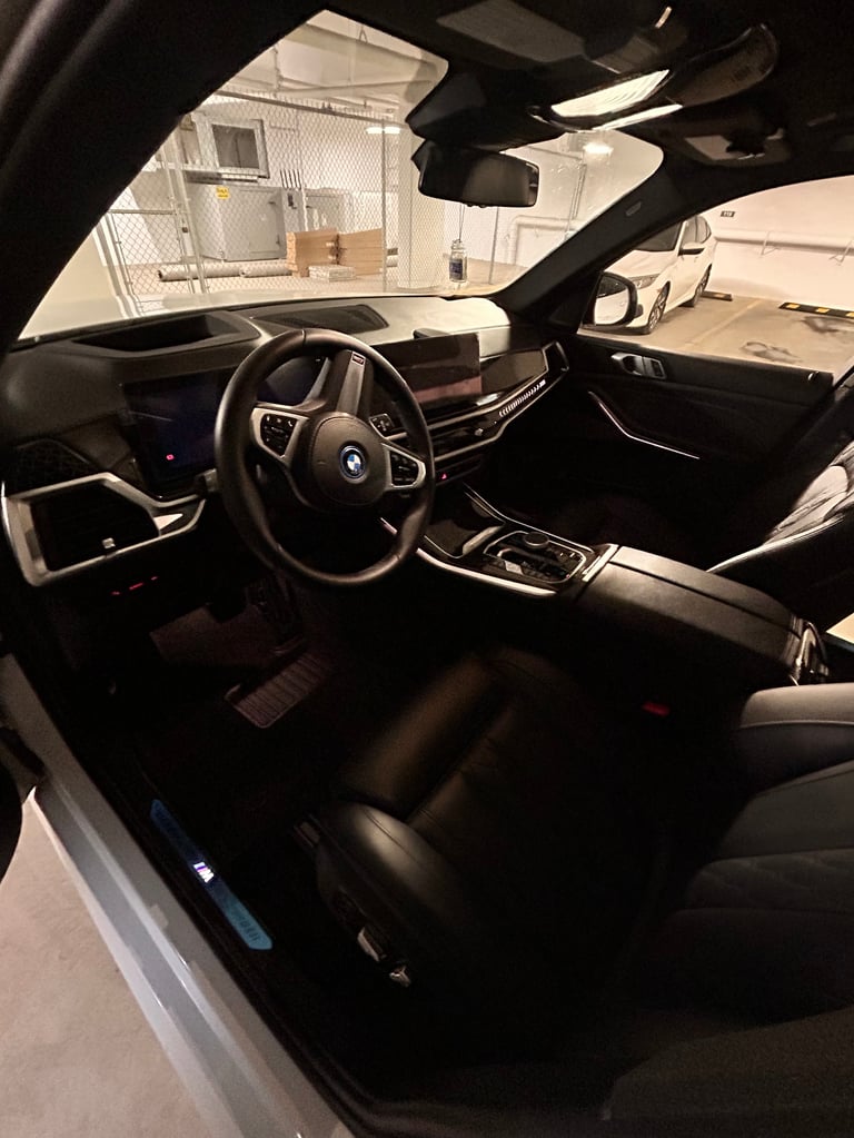 Interior view of a luxury car with dark leather seats, steering wheel, and dashboard, parked in a showroom or garage with white wall visible through windshield