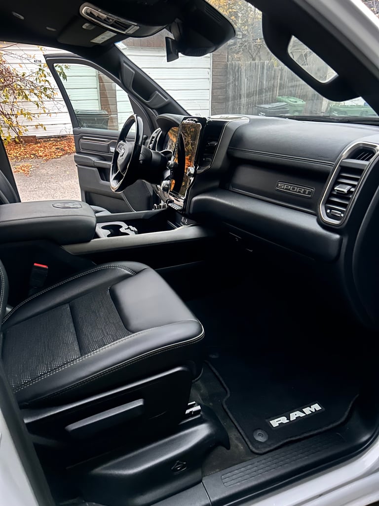 Interior view of a RAM truck showing black leather seats, steering wheel, dashboard, and open driver door with driveway visible outside